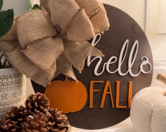 May include: A round wooden sign with the words "hello FALL" in white and orange. A burlap bow is attached to the top left. An orange pumpkin is in the center. Pinecones and a white pumpkin are in the foreground.