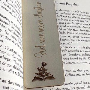 Op de afbeelding: Een houten bladwijzer met een bloemenmotief en de tekst "Just one more chapter" erop gegraveerd. De bladwijzer is lichtbruin en heeft een natuurlijke houtnerf.
