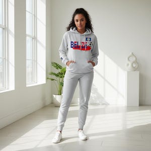 May include: A light gray hoodie and sweatpants set. The hoodie features the word "BELIZE" in blue and red, with a soccer ball graphic. The set includes a matching pair of sweatpants and white sneakers.