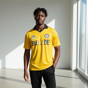 May include: Yellow soccer jersey with black trim and the word "BELIZE" across the chest. The jersey has a v-neck collar and short sleeves. The jersey has a crest on the chest and the sleeves. The person is wearing black pants.
