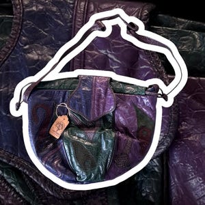 Könnte beinhalten: Vintage Patchwork-Leder-Umhängetasche in Lila- und Grüntönen. Die Tasche hat eine Klappentasche und einen langen Riemen. Ein kleines Etikett hängt an der Tasche. Die Tasche hat ein einzigartiges, strukturiertes Muster.