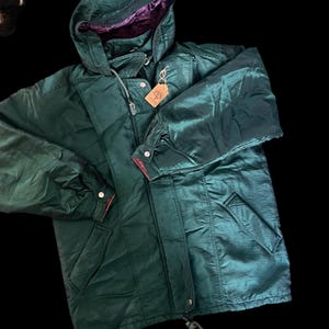May include: A dark green hooded jacket with a shiny, iridescent finish. The jacket has a drawstring hood, a zipper closure, and pockets. The interior lining of the hood is a deep purple color. A small tag hangs from the jacket.