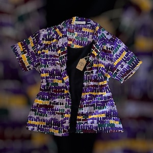 Puede incluir: Una camisa de manga corta con estampado morado, verde y amarillo con una camiseta negra debajo. La camisa tiene cuello y cierre de botones en la parte delantera. La camisa tiene una etiqueta que dice "Prohibition".