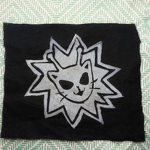 May include: Black fabric patch featuring a stylized cat face within a starburst design. The cat is depicted with large eyes, a small nose, and a smiling mouth. The starburst and cat are in a light gray color.