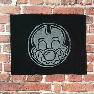 May include: A black fabric patch with a white cartoon character's face. The character has a large nose and a wide smile.