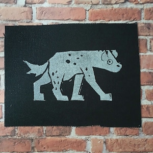 May include: A black fabric patch with a white silhouette of a hyena walking. The hyena has spots and a long tail.