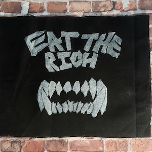 May include: Black fabric patch with the words "Eat the Rich" in white, stylized lettering. Below the words are white, stylized teeth.