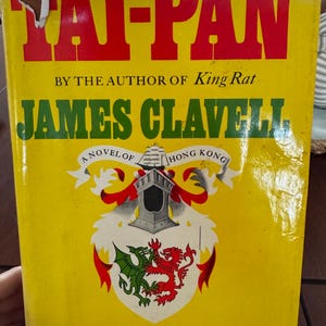 May include: A vintage hardcover book titled "Tai-Pan" by James Clavell. The cover is yellow with red and green text, featuring a coat of arms and the words "A Novel of Hong Kong."
