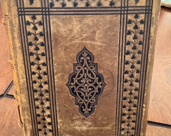 May include: Antique book with a worn, tan leather cover. The cover features a dark blue and black decorative border with floral and geometric patterns. A central, ornate design in dark blue adds detail to the aged book.