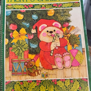 May include: A spiral-bound book titled PACKY'S CHRISTMAS featuring a colorful illustration of a bear in a red outfit holding a candy cane. The cover shows a Christmas tree, wrapped gifts, a small teddy bear, and holly borders in green.