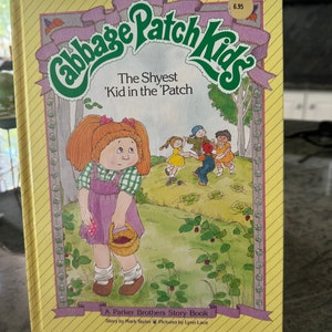 May include: A yellow and purple book with the title "Cabbage Patch Kids" and the subtitle "The Shyest 'Kid in the 'Patch". The book cover features a cartoon illustration of a girl with red hair holding a basket of berries. The book is a Parker Brothers Story Book with a price tag of $6.95.