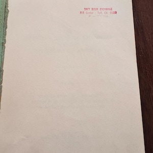 May include: A close-up of a book page with the text "TAFT BOOK EXCHANGE 313 Center - Taft, CA 93268" printed in red. The page is off-white with a green edge and is set against a dark brown surface.