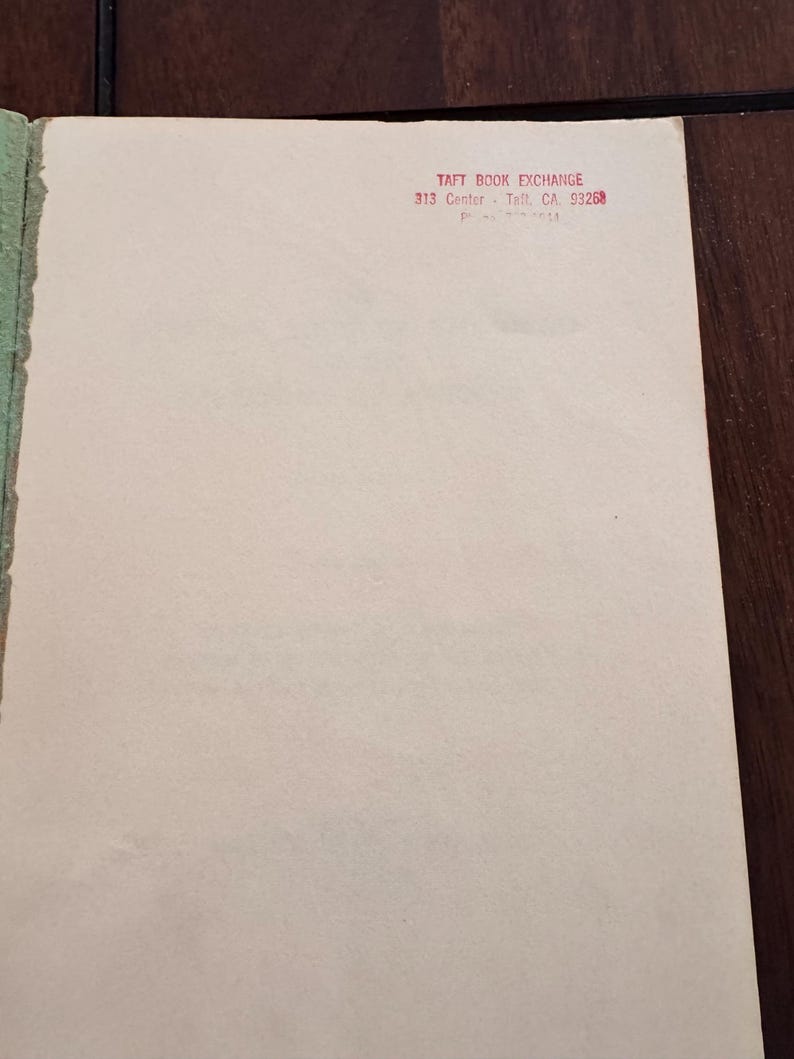 May include: A close-up of a book page with the text "TAFT BOOK EXCHANGE 313 Center - Taft, CA 93268" printed in red. The page is off-white with a green edge and is set against a dark brown surface.