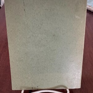 May include: A light green, rectangular object, possibly a book or a piece of art, is propped up on a white, wire stand. The object has a slightly textured surface and appears to be made of paper or cardboard. The background is a dark brown wooden surface.