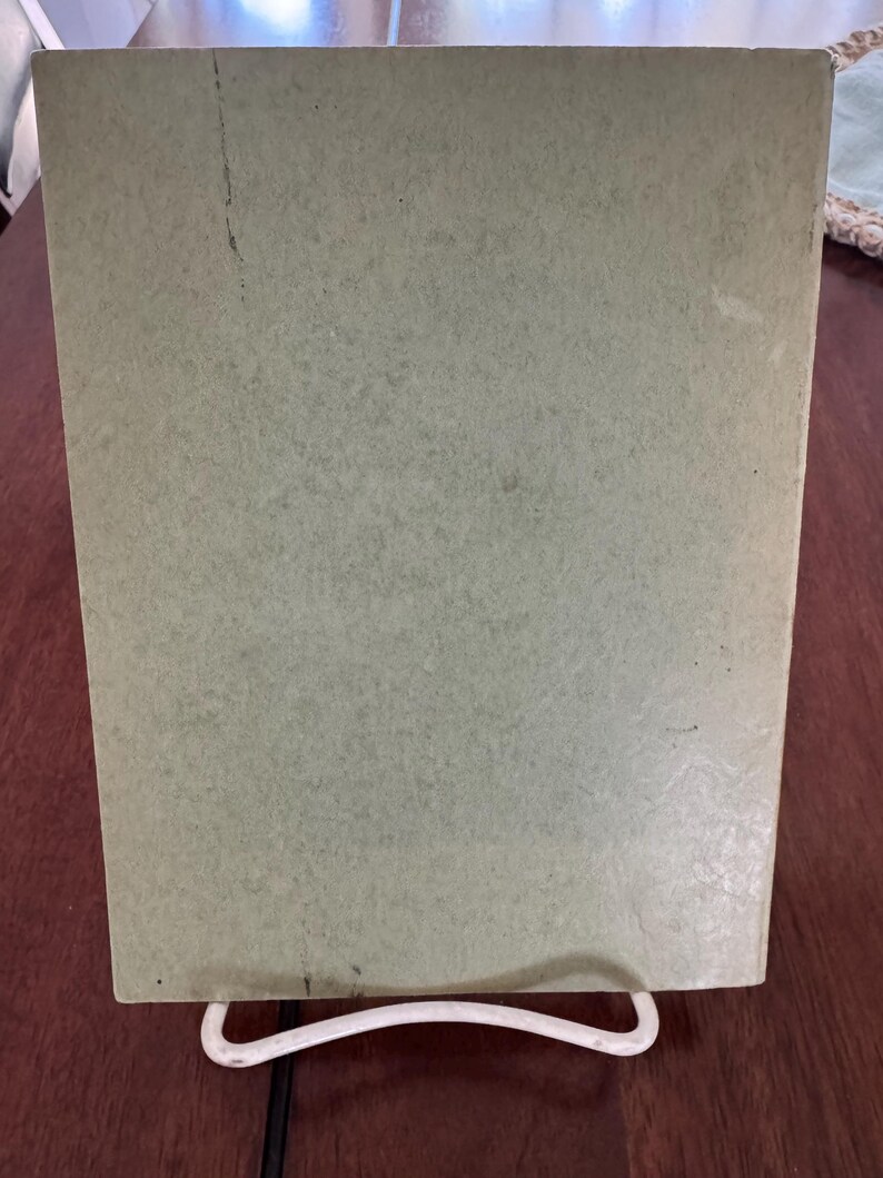 May include: A light green, rectangular object, possibly a book or a piece of art, is propped up on a white, wire stand. The object has a slightly textured surface and appears to be made of paper or cardboard. The background is a dark brown wooden surface.