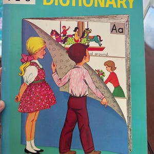 May include: A Young Readers Dictionary for grades 1-2-3 with a teal cover. The illustration shows a girl in a pink floral dress and a boy in a striped shirt looking at a large open book. The book pages feature a carousel and a woman with flowers.