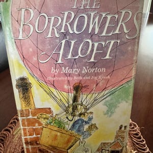 May include: A vintage hardcover book titled "The Borrowers Aloft" by Mary Norton. The cover features an illustration of a hot air balloon with characters and buildings. The book's cover is in shades of pink, red, and white.