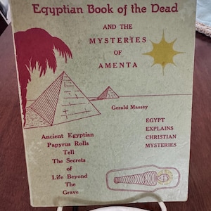 May include: A vintage book titled "The Egyptian Book of the Dead" with red and black text and illustrations. The cover features pyramids, a palm tree, and a sunburst. The book is displayed on a white metal stand.