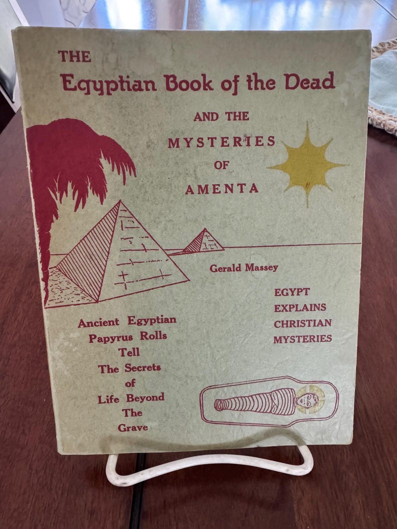 May include: A vintage book titled "The Egyptian Book of the Dead" with red and black text and illustrations. The cover features pyramids, a palm tree, and a sunburst. The book is displayed on a white metal stand.