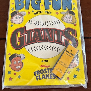 May include: A yellow activity book titled Big Fun With The Giants and Kellogg's Frost Flakes, featuring cartoon characters in baseball caps and a large baseball graphic. A small rectangular box containing four non-toxic crayons is attached.