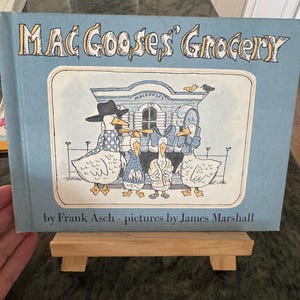 May include: A children's book titled "Mac Goose's Grocery" on a wooden stand. The cover features an illustration of geese in front of a store. The text "by Frank Asch, pictures by James Marshall" is also visible.