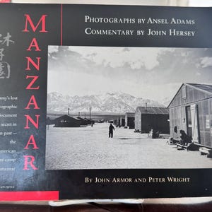 May include: A book cover with the title "MANZANAR" in large red letters. The cover features a black and white photograph of a Japanese-American internment camp. Text includes "Photographs by Ansel Adams" and "Commentary by John Hersey."