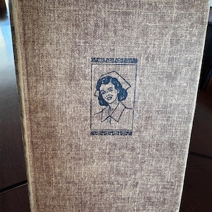 May include: A vintage book with a textured, tan cover. The cover features a black and white illustration of a nurse in a cap, with text above and below the image. The book's spine is visible, and the edges show signs of wear.