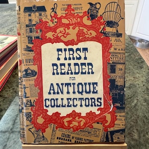 May include: A vintage book titled "First Reader for Antique Collectors" is displayed on a small wooden easel. The book cover is beige with a red and blue decorative frame around the title. The cover features illustrations and text related to antiques.