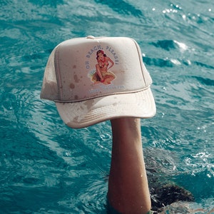 Puede incluir: Una gorra de camionero de malla beige con un gráfico blanco y azul de una mujer en bikini con el texto "Oh Beach, Please!" y "Aloha from Hawaii".