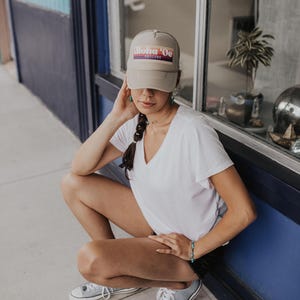 May include: A woman wearing a beige baseball cap with the text "Aloha 'Oe Designs" in a rainbow gradient. She is wearing a white t-shirt and blue jeans. She is sitting on the ground with her legs crossed.