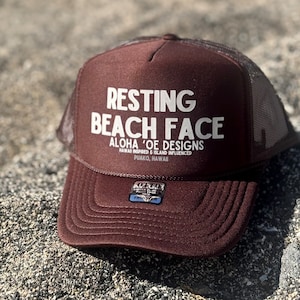 May include: Brown trucker hat with a white mesh back. The front of the hat has white text that reads "Resting Beach Face Aloha 'Oe Designs Hawaii Inspired & Island Influenced Puako, Hawaii"