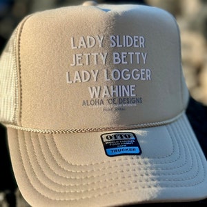 以下が含まれることがあります： ベージュのトラッカーハットに、白い刺繍で「Lady Slider、Jetty Betty、Lady Logger、Wahine、Aloha 'Oe Designs、Island Influenced、Puako、Hawaii」と書いてあります。ハットは黒のメッシュバックで、青いラベルには「Otto、World Famous Foam Front、OSFM、Trucker」と書いてあります。