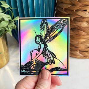 May include: A holographic sticker featuring a black and white illustration of a fairy with wings sitting on a rock. The fairy has long hair and is wearing a dress.