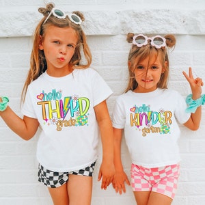 May include: Two young girls wearing white t-shirts with the words "hello THIRD grade" and "hello KINDERgarten" in colorful, retro-style lettering. One girl wears black and white checkered shorts, the other wears pink and white checkered shorts.