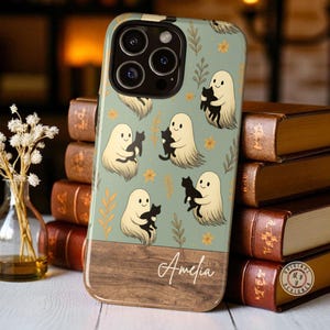 May include: A phone case with a light green pattern of ghosts holding black cats, yellow flowers, and brown leaves. The lower section has a wood grain design with the name "Amelia" in white script.