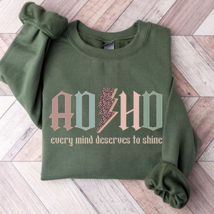 May include: A green sweatshirt with the words "ADHD" in pink, blue, and green letters with a leopard print lightning bolt in the middle. The text "every mind deserves to shine" is below the letters.