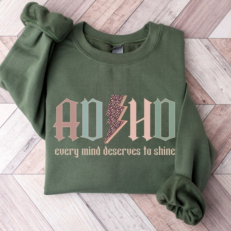 May include: A green sweatshirt with the words "ADHD" in pink, blue, and green letters with a leopard print lightning bolt in the middle. The text "every mind deserves to shine" is below the letters.