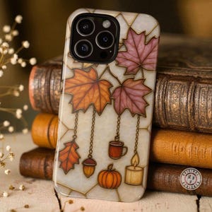 May include: A decorative phone case featuring a fall-themed design. The case has a cream-colored background with a geometric pattern and gold trim. It showcases orange and burgundy leaves, a pumpkin, a mug, an acorn, and a candle.