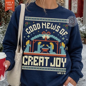 Pode incluir: Uma camisola azul-marinho de gola redonda com uma estampa gráfica de uma cena de natividade com o texto "GOOD NEWS OF GREAT JOY" e "LUKE 2:10" num padrão de estilo malha amarelo, azul-claro e vermelho no peito.