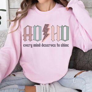 May include: Pink sweatshirt with the text "ADHD" in pastel colors with a leopard print lightning bolt in the middle. The text "every mind deserves to shine" is below the graphic.