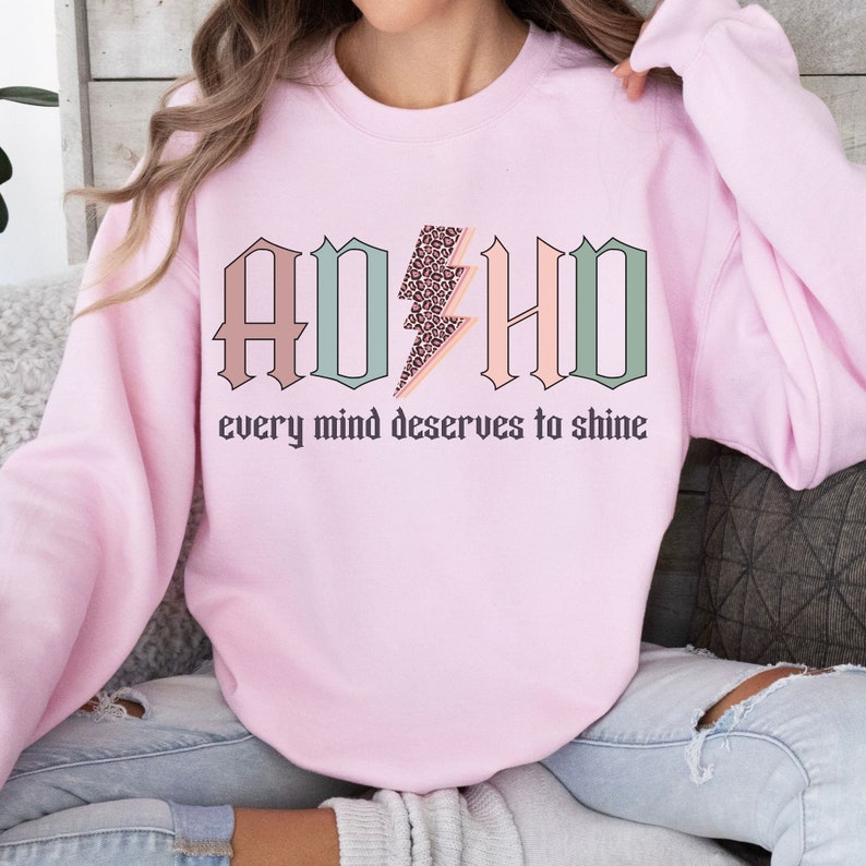 May include: Pink sweatshirt with the text "ADHD" in pastel colors with a leopard print lightning bolt in the middle. The text "every mind deserves to shine" is below the graphic.