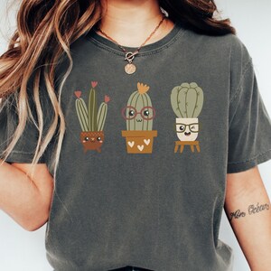 May include: A dark grey t-shirt featuring a graphic of three cartoon cacti in pots. The cacti are wearing glasses and have smiling faces. The text "More Ocean" is written on the right sleeve of the shirt.