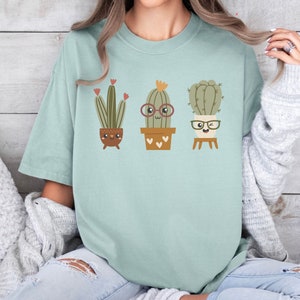 May include: A light blue t-shirt featuring a graphic of three cartoon cacti wearing glasses. The cacti are in pots with hearts on them.