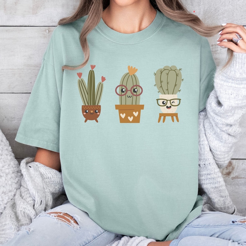 May include: A light blue t-shirt featuring a graphic of three cartoon cacti wearing glasses. The cacti are in pots with hearts on them.