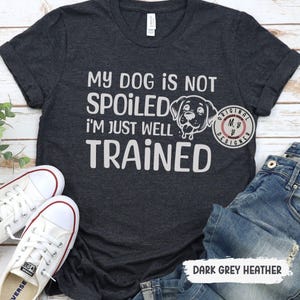 May include: Dark grey heather t-shirt with the text "My dog is not spoiled I'm just well trained" and a cartoon image of a dog. The text "Original MB Designer" is in a circle in the top right corner of the shirt.