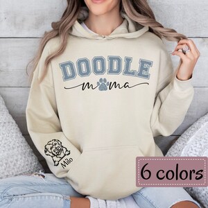 May include: A beige hoodie with the text "Doodle mama" in blue with a paw print. The left sleeve has a small doodle dog illustration with the name "Milo" underneath.