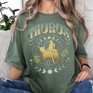 May include: Green t-shirt with a vintage-style graphic of a woman riding a bull with the word "TAURUS" above it.