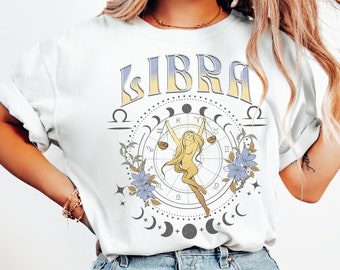 May include: White t-shirt with a graphic design featuring the word "LIBRA" in a vintage font, a stylized image of a woman holding a scale, and a circle of astrological symbols.