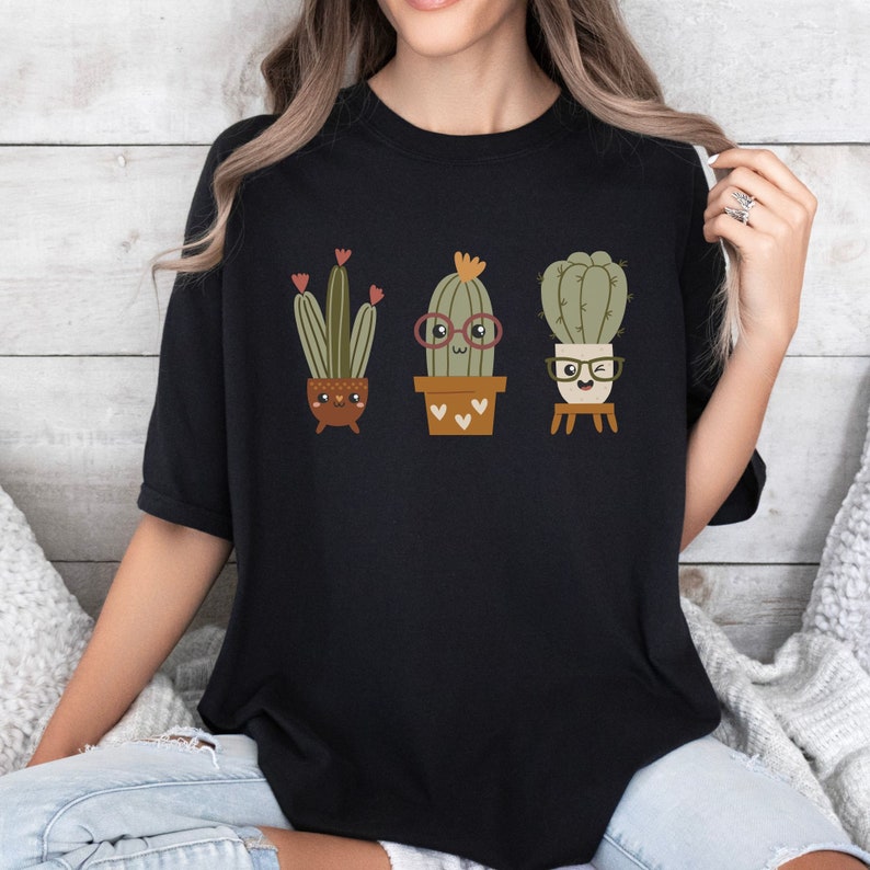 May include: Black t-shirt featuring a graphic of three cartoon cacti wearing glasses and smiling. The cacti are in pots with hearts on them.