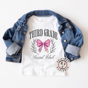 May include: White t-shirt with the text "Third Grade Social Club" in black lettering. A pink bow with a pencil through the center is above the text.